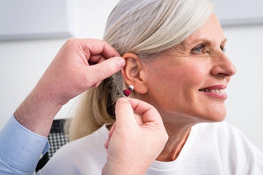 Seniorin bekommt modernes Hörgerät vom Hörakustiker ins Ohr eingesetzt und lächelt.